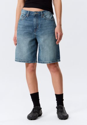 Person iført blå denimshorts i mellemlang længde, sorte ankelsokker, sorte teksturerede sko og sort kort top, stående foran en ensfarvet baggrund.