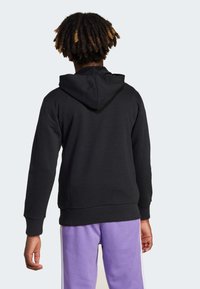 Schwarze Kapuzenjacke mit Kängurutasche und Kordelzugkapuze, kombiniert mit lila Hosen mit weißen Seitenstreifen. Strukturierter Baumwollstoff.