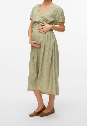 Personne enceinte portant une robe midi à fleurs vert clair et des ballerines marron, les mains caressant doucement son ventre arrondi sur un fond blanc.