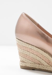 Scarpa con zeppa caratterizzata da una tomaia in rosa gold metallizzato, con tacco in corda jute testurizzata, arricchita da fili rosa intrecciati nel design.