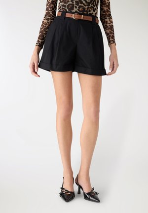 Donna che indossa pantaloncini neri a vita alta, top a maniche lunghe con stampa leopardata, cintura marrone e scarpe nere con tacco a punta, in piedi su sfondo bianco.