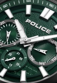 Žalias tekstūruotas laikrodžio ciferblatas su sidabriniais valandų žymekliais, dvigubais subdijalu, skirtais dienai ir datai, ir išsiskiriančiu "POLICE" logotipu.