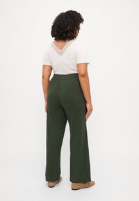 Pantalones verde de pierna ancha con un acabado texturizado, combinados con una blusa blanca que presenta detalles de encaje en la parte posterior del cuello. Botines tobilleros color beige claro.