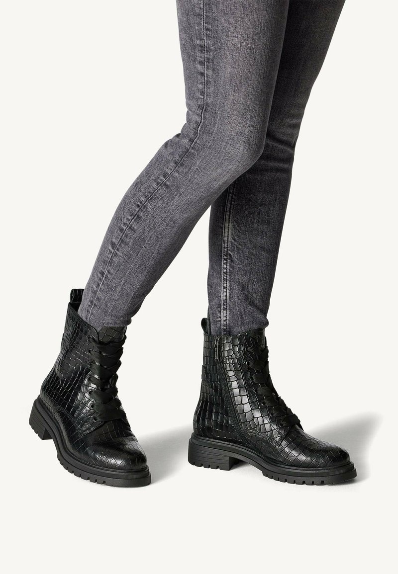 Bottines noires en cuir texturé à lacets avec semelles épaisses portées avec un jean gris ajusté sur un fond uni.