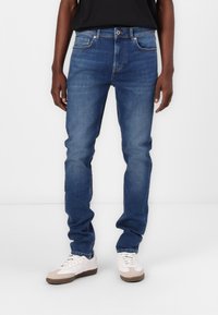 Blue denim jeans with a slim fit, five-pocket design, and subtle stitching details. Paired with white and light brown sneakers.
