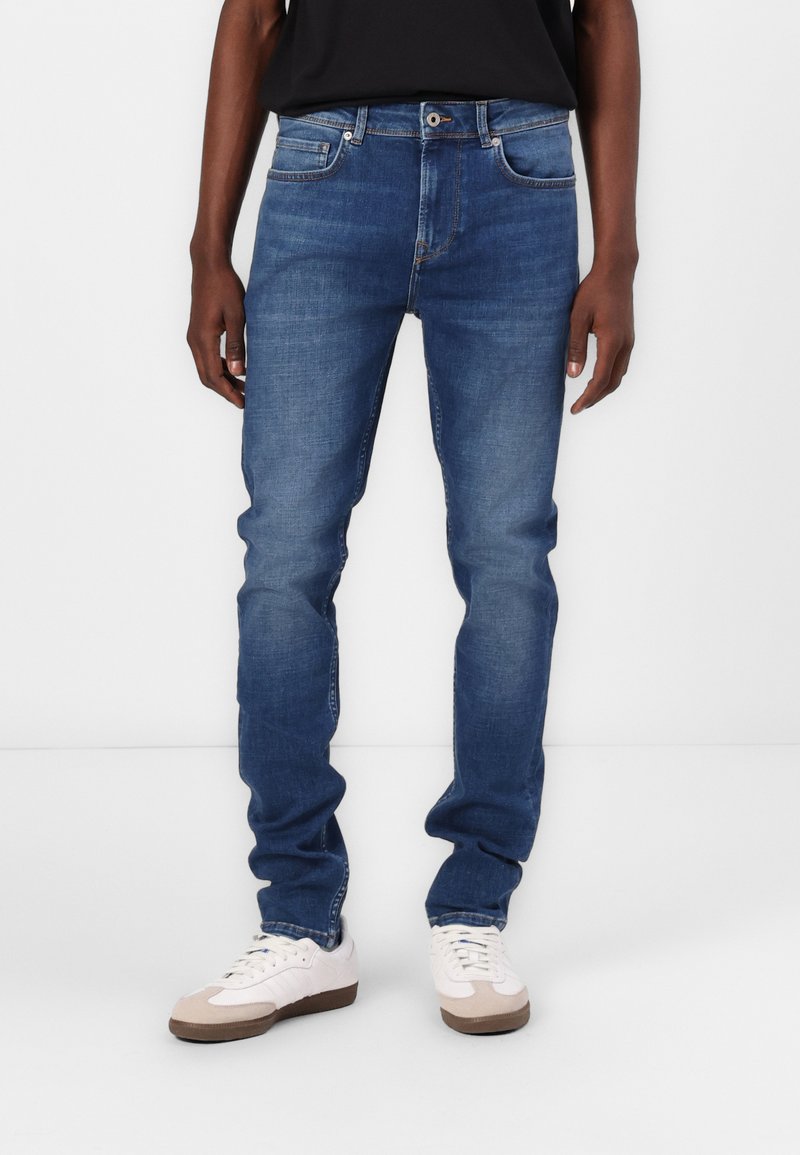 Jeans de mezclilla azul con un corte slim, diseño de cinco bolsillos y detalles de costura sutiles. Combinados con zapatillas blancas y marrones claros.