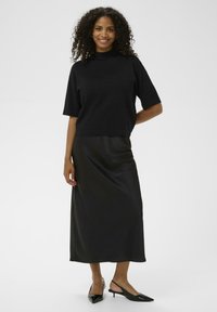 Zwarte top met korte mouwen en een hoge halslijn, gecombineerd met een aansluitende zwarte satijnen maxirok. Model draagt zwarte slingback schoenen met een spitse neus.