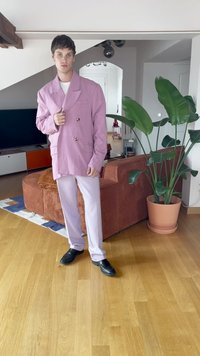 Giovane uomo in piedi in un soggiorno, indossa un blazer oversize viola pastello, pantaloni abbinati e scarpe nere, vicino a un divano marrone e a una grande pianta verde.