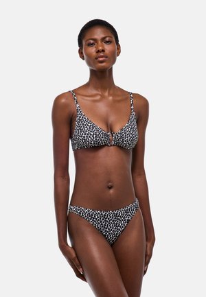 Femme debout, portant un bikini imprimé léopard noir et blanc avec une découpe en forme d'œillet sur le haut, sur un fond clair uni.