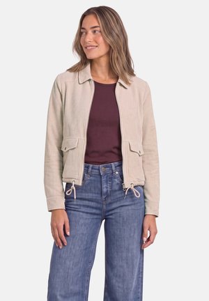 Femme portant une veste en velours côtelé beige sur une chemise rouge foncé et un jean bleu, debout devant un fond blanc, regardant sur le côté et souriant.