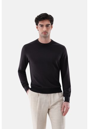 Mann trägt ein schwarzes Langarmshirt und beige Hosen, steht mit einer Hand in der Tasche vor einem schlichten weißen Hintergrund.