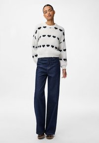 Vit stickad tröja med svarta hjärtan, kombinerad med högmidjade mörkblå vida jeans och bruna skor. Slät textur överlag.