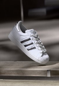 Baskets Adidas blanches avec des rayures noires et un bout en coque, positionnées en plein air au-dessus d'une surface en béton dans un éclairage doux.
