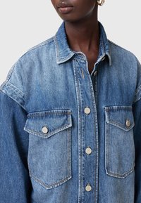 Denim jaka ar divām krūšu kabatām, sudraba pogu aizdares un izbalējušo zilu gradientu. Tekstūrēta auduma ar redzamām šuvēm.