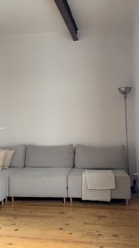 Graues Ecksofa mit Kissen und einer gefalteten beigen Decke, Holzboden, hohe silberne Stehlampe und schlichte weiße Wand mit einem Deckenbalken.