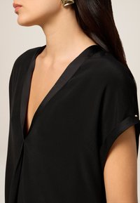 Blusa nera con scollo a V e maniche corte, realizzata in un materiale liscio, con rifiniture in satin lungo il collo e i bordi delle maniche.