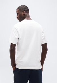 Άνδρας με κοντά μαλλιά που φοράει απλό λευκό μπλουζάκι και σκούρο παντελόνι, γυρισμένος πλάτη με τα χέρια στις τσέπες, πάνω σε ανοιχτό γκρι φόντο.