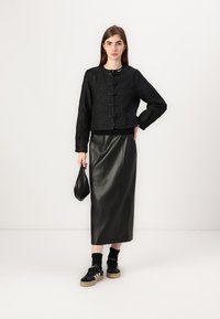 Black cropped jacket with textured fabric and front clasp, paired with a black leather midi skirt and black sneakers. Holds a small black bag.