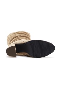 Beige suède enkellaars met een gerimpeld ontwerp en een blokhak. De zool is van zwart rubber met een gestructureerd patroon voor grip.