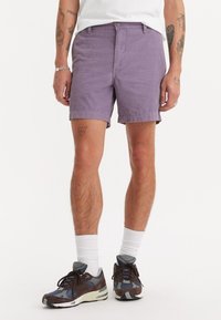 Pantaloni scurți mov din material, cu un talie standard, buzunare laterale și un tiv deasupra genunchiului. Purtați cu șosete albe și pantofi sport.