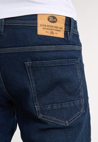 Jeans de mezclilla azul oscuro con un parche de cuero con la marca "Petrol", detalles visibles en el bolsillo trasero y costuras reforzadas.