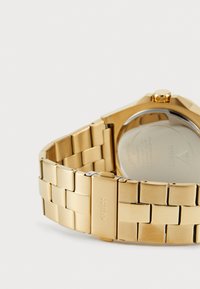Guldtonad klocka med en metalllänkarmband, i kvadratisk design och med en genomskinlig baksidan. Stängning med "GUESS" gravyr.
