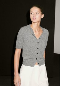 Cardigan a maniche corte con bottoni di colore grigio, in maglina strutturata, con rever a scollo e bottoni neri, indossato sopra pantaloni chiari.