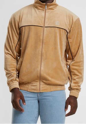 Mand iført en beige velourjakke med lynlås og sorte kantbånd samt lyseblå jeans, stående med hænderne afslappet ned langs siden.