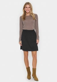Chemise longue à manches marron associée à une jupe noire en A. Le mannequin porte des bottines marron. Design simple avec un tissu texturé sur la jupe.