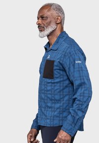 Blaues kariertes Hemd mit einer schwarzen Brusttasche, Knopfverschluss und langen Ärmeln mit Manschettenknöpfen. Der Stoff hat eine glatte Textur.