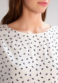 Blusa bianca caratterizzata da un delicato motivo di piccole foglie blu navy. Il tessuto è leggero, con una superficie liscia e una scollatura rotonda.