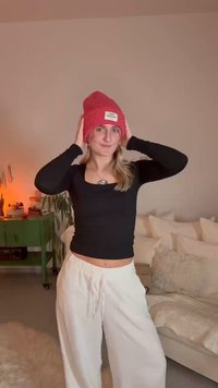 Κόκκινο ριγωτό σκουφί με ετικέτα, μαύρο μακρυμάνικο τοπ και άσπρο άνετο παντελόνι. Το φόντο απεικονίζει έναν άνετο εσωτερικό χώρο με απαλές υφές.