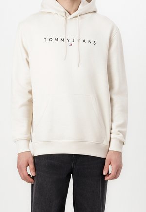 Mężczyzna noszący kremową bluzę z kapturem z napisem "TOMMY JEANS" i małym logo na środku klatki piersiowej, w połączeniu z czarnymi dżinsami.
