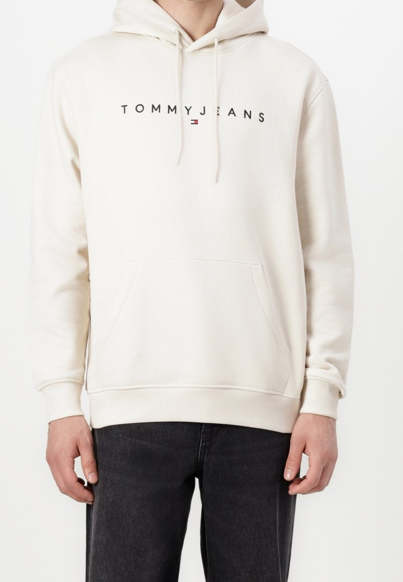 Homme portant un sweat à capuche blanc cassé avec le texte "TOMMY JEANS" et un petit logo centré sur la poitrine, associé à un jean noir.