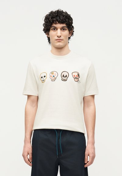 Jeune homme aux cheveux bouclés et foncés portant un t-shirt blanc avec quatre illustrations colorées de crânes sur la poitrine et un short foncé avec des cordons bleus.