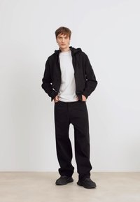 Sudadera negra con cremallera sobre una camiseta blanca, combinada con pantalones negros holgados y zapatillas negras; tejido suave, ajuste casual, diseño minimalista.