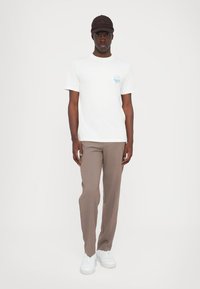 Hvid t-shirt med et blåt grafik på brystet, parret med brune bukser og hvide sneakers. Modellen har også en brun kasket på.