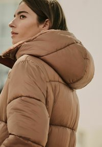 Chaqueta de plumas marrón con cuello alto, forro interior suave y textura acolchada. El material parece ligero pero cálido.