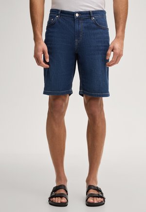 Mann trägt kniehohe dunkelblaue Jeansshorts und schwarze Sandalen mit zwei Riemen, steht vor einem einfarbigen Hintergrund.