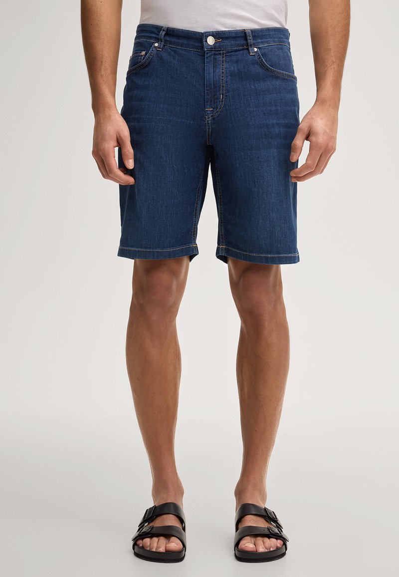 Mann trägt kniehohe dunkelblaue Jeansshorts und schwarze Sandalen mit zwei Riemen, steht vor einem einfarbigen Hintergrund.