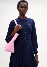 Abito navy a maniche lunghe con colletto e bottoni frontali, dotato di una cintura con fiocco. Una borsa rosa texturizzata è tenuta con una mano.