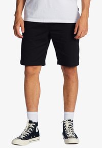 Pantaloni corti neri in cotone con una vestibilità dritta, tasche laterali e un orlo che arriva appena sopra il ginocchio, abbinati a scarpe sportive alte nere.