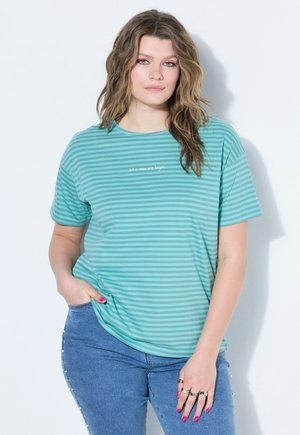 Vrouw met golvend haar draagt een T-shirt met turquoise en groene strepen met de tekst "laat een nieuw tijdperk beginnen", blauwe jeans met pareldetails op de zijnaden.