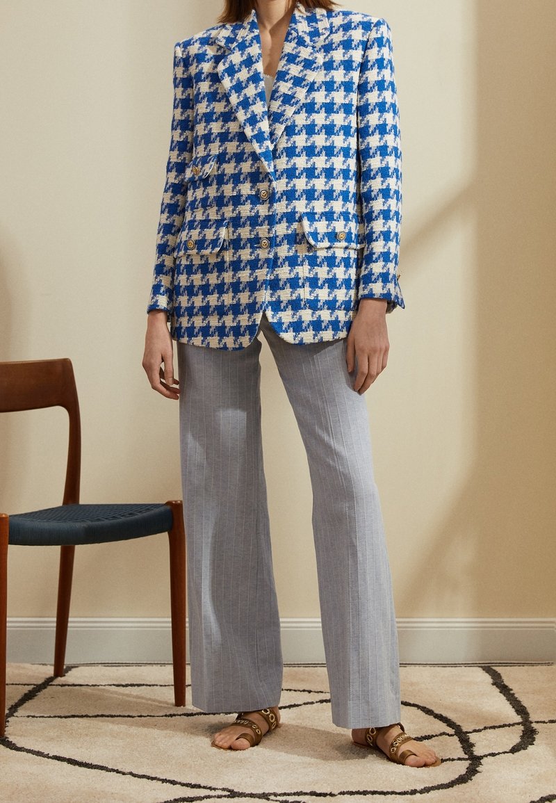 Femme portant un blazer pied-de-poule bleu et blanc, un pantalon large gris clair à fines rayures et des sandales plates marron, debout à l'intérieur près d'une chaise en bois.