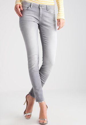 Kobieta ubrana w jasnoszare obcisłe dżinsy, żółto-białą bluzkę w paski z długim rękawem oraz srebrne sandały na wysokim obcasie, stojąca ze skrzyżowanymi nogami.