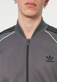 Siva jakna na zip s uspravnim ovratnikom, crnim Adidas logotipom i tri crne pruge na ramenima. Mekana, glatka tekstura.