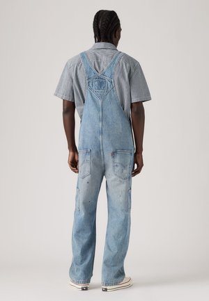 Person mit geflochtenem Haar steht mit dem Rücken zur Kamera, trägt hellblaue Denim-Latzhosen, ein kurzärmeliges gestreiftes Shirt und weiße Turnschuhe.