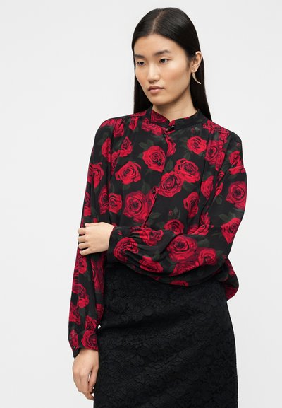 Μαύρη μπλούζα με κόκκινα σχέδια τριαντάφυλλου, μακριά μανίκια και κλείσιμο με κουμπιά. Φορεμένη πάνω από μαύρη φούστα από δαντέλα, με υφασμένη λεπτομέρεια.