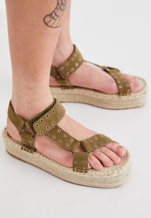 Pieds chaussés de sandales compensées en daim vert olive avec des œillets et des semelles en jute tissé, avec un tatouage partiel visible sur la jambe.