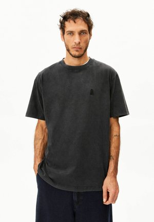 Mann mit kurzen lockigen Haaren und Bart trägt ein weites schwarzes gewaschenes T-Shirt und dunkle Hosen, steht mit einer Hand in der Tasche vor weißem Hintergrund.